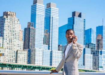 Young African American Businessman traveling in New York City, wearing patterned blazer, white shirt, standing in business district with high buildings of Manhattan under sun, talking on cell phone."n