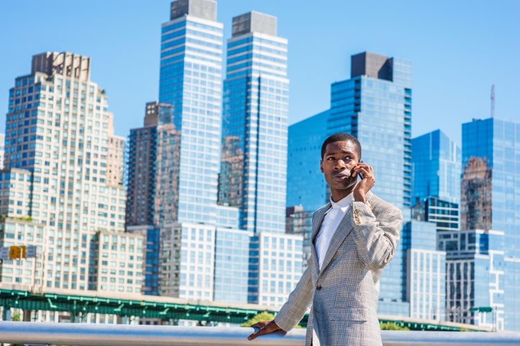 Young African American Businessman traveling in New York City, wearing patterned blazer, white shirt, standing in business district with high buildings of Manhattan under sun, talking on cell phone."n