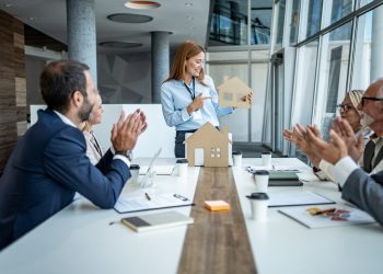 Real estate agent presenting a cardboard house model to colleagues, who are applauding enthusiastically in a collaborative office meeting, celebrating teamwork and innovative design achievements