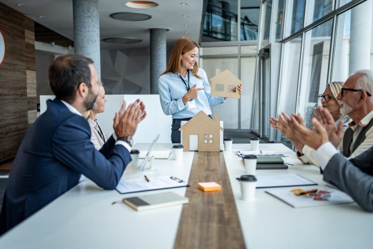 Real estate agent presenting a cardboard house model to colleagues, who are applauding enthusiastically in a collaborative office meeting, celebrating teamwork and innovative design achievements